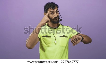 Man referee checking his watch and pointing finger, whistle on lanyard and headset mic visible, yellow referee jersey against purple studio wall; urgency.