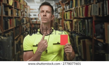 Man referee points finger at red card while holding whistle wearing neon yellow jersey in library building, eyes closed; authority.
