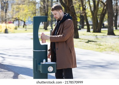 Man refilling his water bottle at the city. Free public water bottle refill station. Sustainable and green city. Male in brown coat.  Tap water to reduce plastic bottle usage. Drinking water dispenser