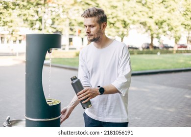 Man refilling his water bottle at the city. Free public water bottle refill station. Sustainable and green city. Male in white shirt. Tap water to reduce plastic bottle usage. Drinking water dispenser
