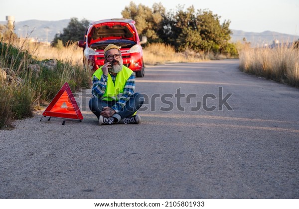 Man with relaxed attitude\
stranded on the road next to the broken down car talking on the\
phone