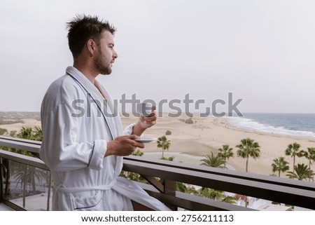 A man relaxes in a bathrobe while sipping coffee on his balcony, with breathtaking views of the Gran Canaria beach and ocean. The perfect start to a luxurious holiday.