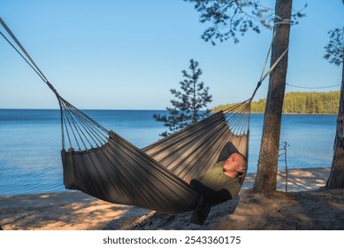 A man relaxes in a hammock by a forest lake, enjoying a serene view of the sandy shore, clear blue sky, and surrounding trees. Capturing the essence of tranquility and nature's peaceful retreat - Powered by Shutterstock