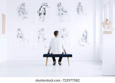 Man relaxing in art gallery while sitting on stool in front of white wall with drawings. Art gallery concept