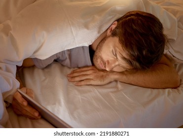 Man Relaxing In Bed On A Digital Tablet Browsing On Social Media Or Networking On The Internet. Male Addicted To Technology Scrolling On Mobile At Night In A Room. Guy Searching The Web With An Ipad