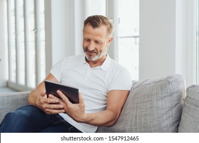 Man Relaxing At Home On A Comfortable Sofa With His Tablet Pc Reading Online With A Quiet Smile Of Pleasure