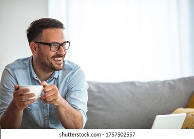 Man relaxing at home on the couch and having a coffee break, he is smiling and holding a cup. Handsome man relaxing with cup of coffee at home. The day doesn't start until that first sip