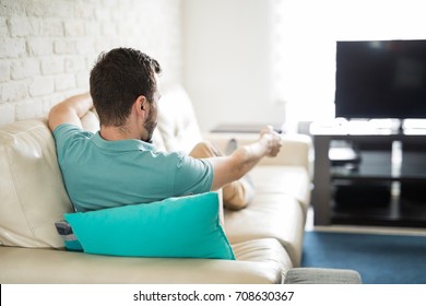 Man Relaxing At Home Sitting In His Cozy Sofa Holding A Remote Control In His Hand And Deciding What To Watch On Tv