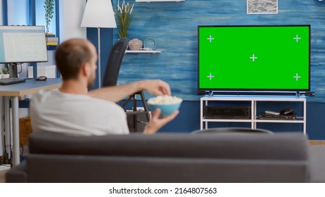 Man relaxing on couch looking at green screen on tv watching movie while eating popcorn in modern living room. Person having a good time sitting on sofa looking at television with chroma key display. - Shutterstock ID 2164807563