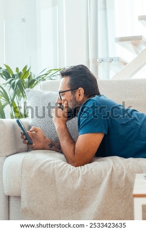 Man relaxing on the couch while using his smartphone to explore social media, watching videos, and connecting with friends, representing a contemporary lifestyle of leisure and digital engagement