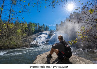 Man Relaxing On Hiking Trip. Hiker With Backpack Sitting On The Rock Reading Tablet By The Waterfall. High Falls  Of Dupont State Forest. Near Asheville, Western North Carolina, USA. 