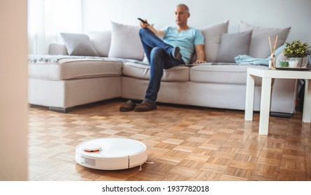 Man Relaxing On Sofa With Robotic Vacuum Cleaner On Hardwood Floor. Cleaning concept - automatic robotic hoover clean the room while man relaxing, close up