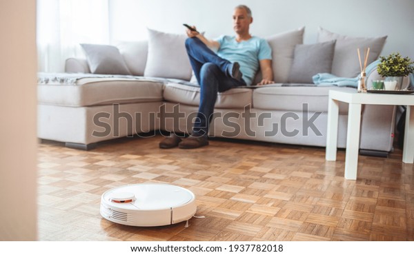 Man Relaxing On Sofa With Robotic\
Vacuum Cleaner On Hardwood Floor. Cleaning concept - automatic\
robotic hoover clean the room while man relaxing, close\
up