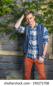 Man Relaxing Outside. Wearing A Blue And White Pattern Shirt,  A Blue Hoody Vest,  Red Jeans, One Hand Scratching Head,  Young Guy Is Standing By A Wall With Green Ivy Leaves, Relaxing And Thinking