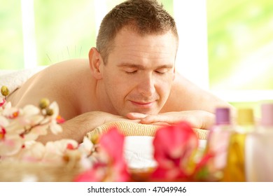 Man relaxing in sunny salon with closed eyes laying on the mat with acupuncture needles on back as alternative therapy with flowers and cosmetics