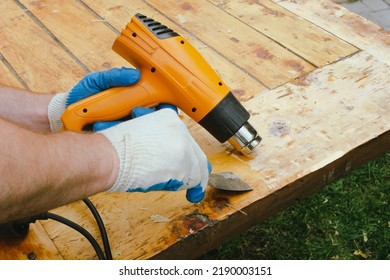 Man Removing Old Varnish And Paint From Wood Using Scraper And Heat Gun. Restoration Work Of Old Wooden Table Outdoors. Furniture Repair, Reuse Of Old Things Concept.