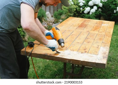 Man Removing Old Varnish And Paint From Wood Using Scraper And Heat Gun. Restoration Work Of Old Wooden Table Outdoors. Furniture Repair, Reuse Of Old Things Concept.