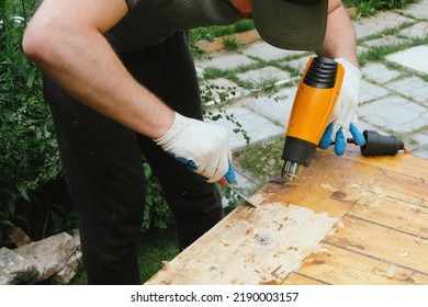 Man Removing Old Varnish And Paint From Wood Using Scraper And Heat Gun. Restoration Work Of Old Wooden Table Outdoors. Furniture Repair, Reuse Of Old Things Concept.