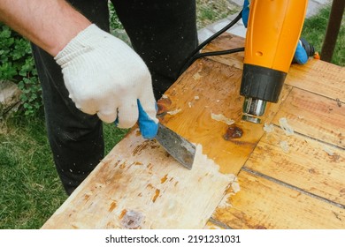 Man Removing Old Varnish And Paint From Wood Using Scraper And Heat Gun. Restoration Work Of Old Wooden Table Outdoors. Furniture Repair, Reuse Of Old Things Concept.