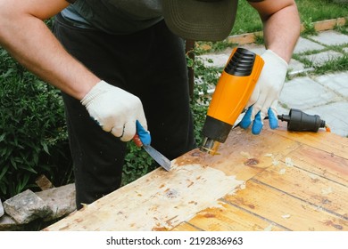 Man Removing Old Varnish And Paint From Wood Using Scraper And Heat Gun. Restoration Work Of Old Wooden Table Outdoors. Furniture Repair, Reuse Of Old Things Concept.