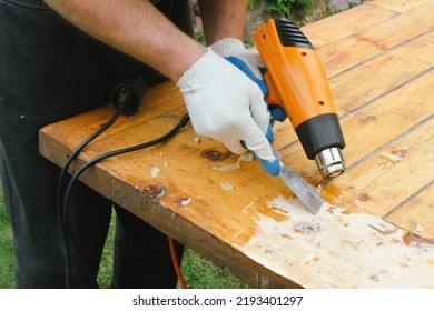 Man Removing Old Varnish And Paint From Wood Using Scraper And Heat Gun. Restoration Work Of Old Wooden Table Outdoors. Furniture Repair, Reuse Of Old Things Concept.