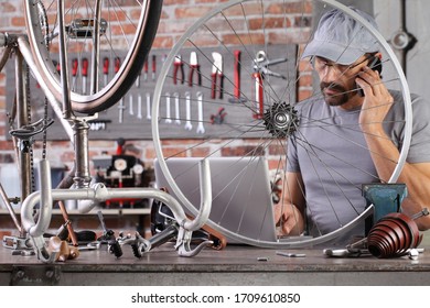 Man Repair The Vintage Bicycle With Mobile Phone At The Computer Search Spare Parts On The Internet In Garage Workshop On The Workbench With Tools, Diy Concept