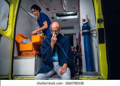 Man Rescued From The Fire Sitting In An Ambulance Car, Breathing Through An Oxygen Mask, A Female Paramedic In The Background.