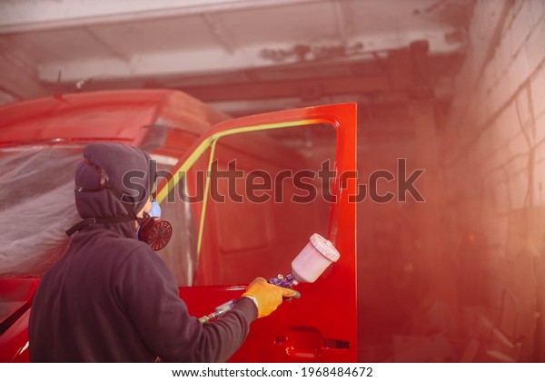 man in a respirator paints a car with a spray\
gun in red, repair a truck\
body.