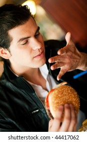 Man In A Restaurant Or Diner Eating A Hamburger, It Is So Good He Is Licking His Fingers, Shot With Available Light, Very Selective Focus