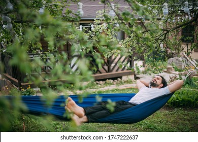 A man is resting in a hammock in a country house. A young guy lies in a hammock, dozing outdoors in the summer in the green of trees in the country - Shutterstock ID 1747222637