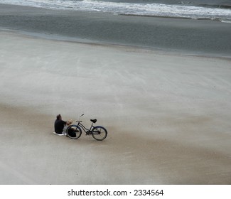 Man resting on an empty beach overlooking the ocean on an overcast day.  A bicycle lays by his side.