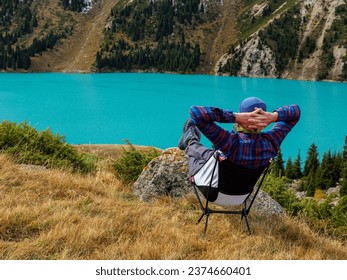 A man is resting while sitting on a folding chair in the mountains on the shore of a beautiful lake. Enjoying a magnificent panoramic view of snow-capped peaks and a turquoise mountain lake.