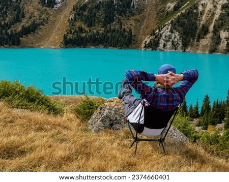 A man is resting while sitting on a folding chair in the mountains on the shore of a beautiful lake. Enjoying a magnificent panoramic view of snow-capped peaks and a turquoise mountain lake.