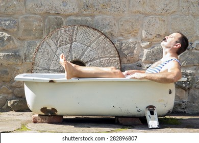 Man In Retro Swimsuit Resting In The Outdoor Bathtub. Man In The Old Outdoor Tub A Sunbathe Face.  Recreational Relaxation In A Swimsuit At The House.