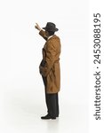 Man in retro-style trench coat and fedora hat standing and pointing upwards isolate don white background. Concept of retro and vintage, fashion, business, detective