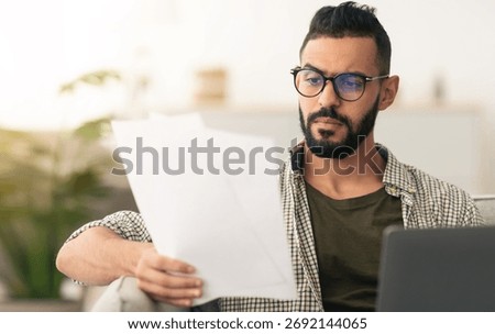 Man Reviews Documents While Sitting on a Couch Near a Laptop in a Modern Living Space
