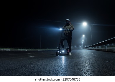 A man rider on an electric scooter is standing on the road, a foggy autumn night, the light from the lanterns