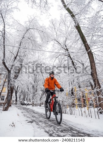 A man rides a bicycle through a winter city among snow covered trees. The guy on the red bike is wearing an orange jacket and a bicycle helmet on his head. Active lifestyle in winter
