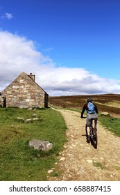 A Man Riding A Bicycle On A Lonely Road In Scottish Highlands Past A Small Stone House Or Shelter.  