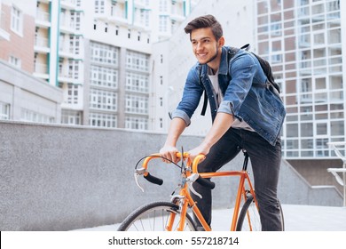 Man Riding A Bicycle Outside. Attractive Young Boy In Blue Jeans Jacket With White Shirt Underneath Cycling With Great Joy To His Friends. Urban Background