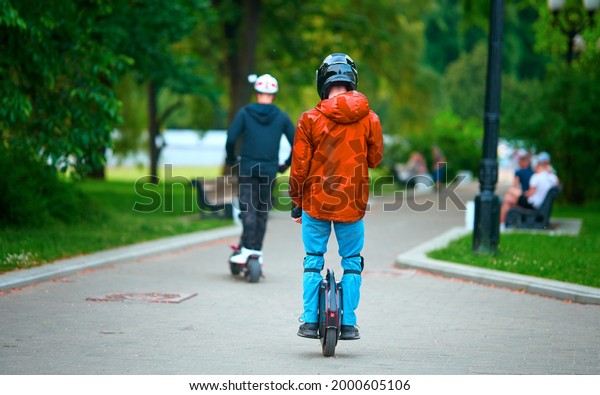 Man riding on unicycle in protective gear\
through public park. Young man with helmet and protective equipment\
rides on electric mono wheel in park on summer day. Urban\
individual transport