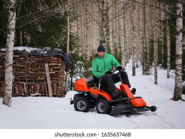 Man riding orange lawn mower in snow with birch forest and stacked firewood in background