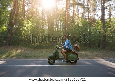 Man riding scooter on asphalt road in green sunny forest. Concept of travelling, vacation, journey and trip. Side view of young caucasian guy in glasses