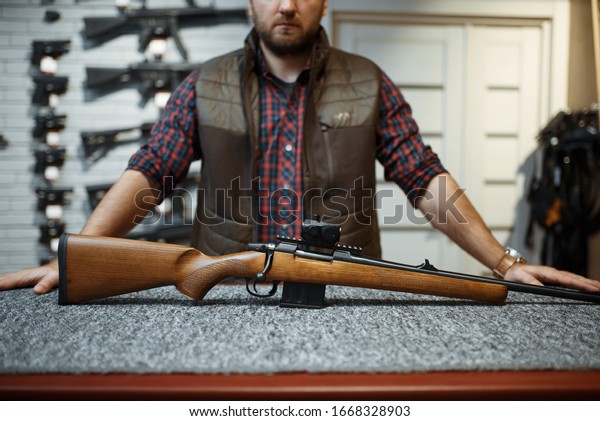Man with rifle\
standing at counter in gun\
shop