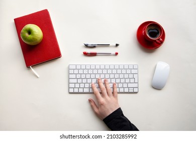 Man right hand on wireless keyboard with red notebook, green apple, cup of coffee, mouse, black, red pens on white table