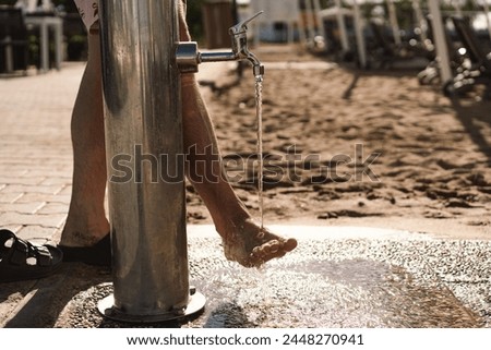 A man rinses his bare feet under a stainless steel faucet, the drops glistening in the sun. An urban environment with a sandy area indicating the likely proximity to a beach where people can cool off