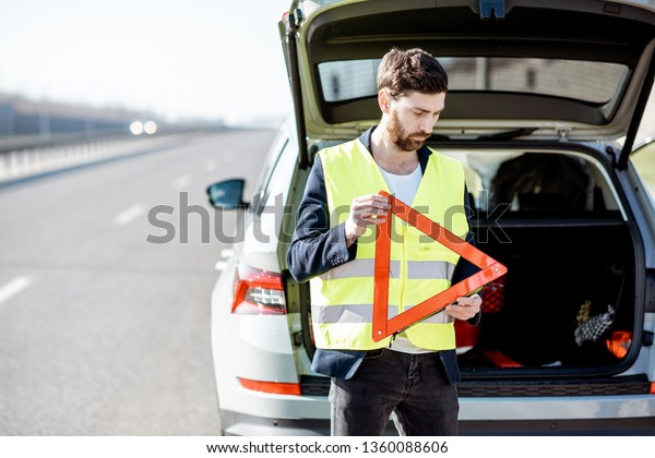 Man in road vest holding emergency triangle\
sign near the broken car on the\
roadside