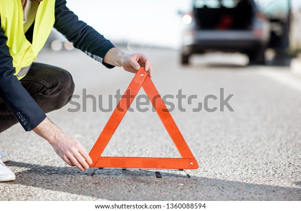 Man in road\
vest putting emergency triangle sign on the highway with broken car\
on the background, close-up\
view