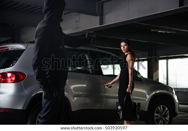 Man robber in black hoodie threatening\
with gun to scared businesswoman on car\
parking