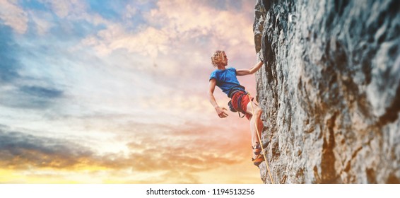 man rock climber with long hair. side view of young man rock climber in bright red shorts resting while climbing the challenging route on the cliff on the colorful down sky background. rock climber